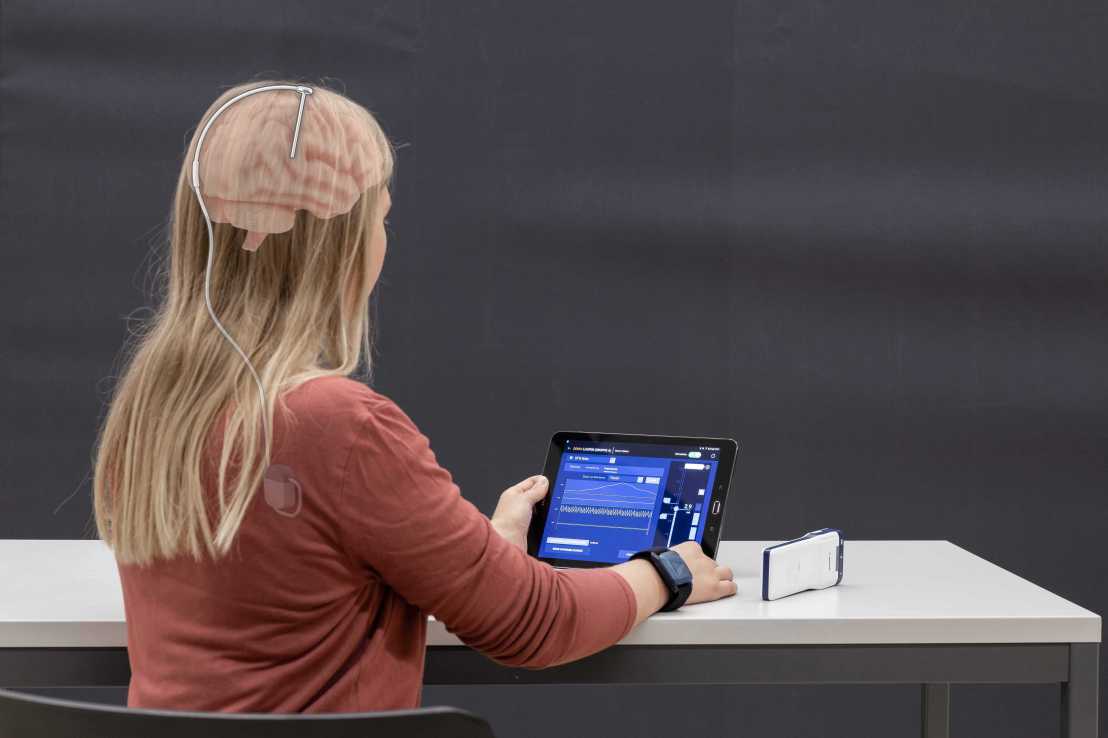 Enlarged view: Woman photographed from behind, holding a tablet. Overlaid with a graphic showing her brain, where a sensor is installed to measure  brain function.