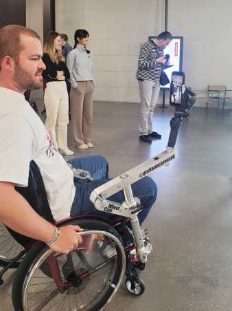 Enlarged view: Side-view of a student sitting in a manual wheelchair with Giuliano’s camera mount and a smartphone attached