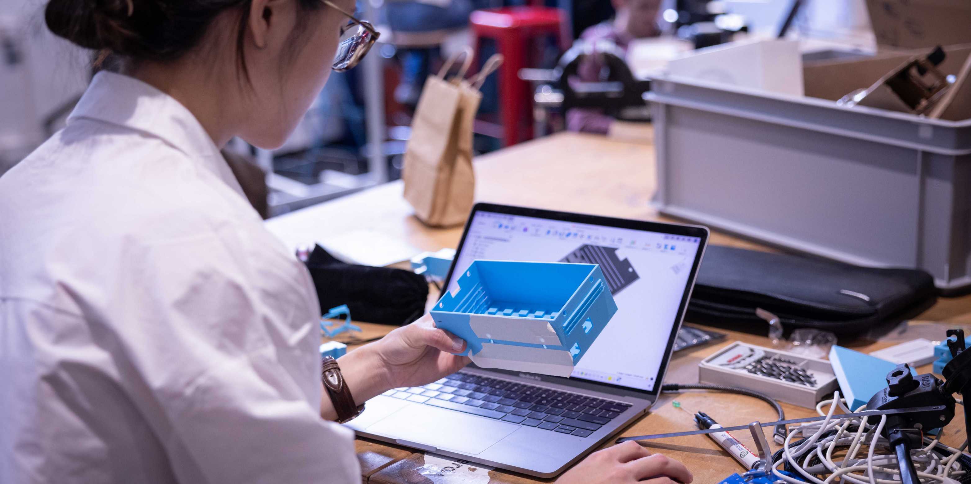 Student holding a blue 3D-printed box, working on a laptop on a CAD model.