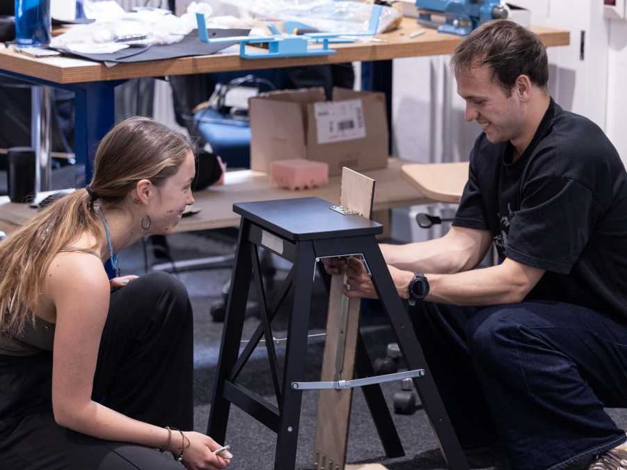 Enlarged view: Two students kneeling in front of a prototype: a black chair with a wooden backrest attached.