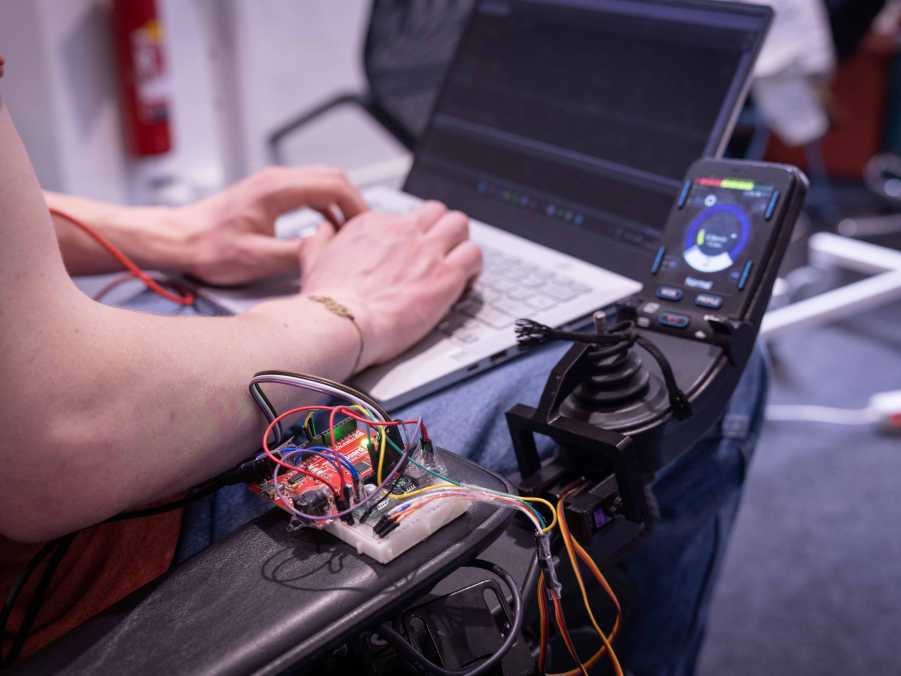 Enlarged view: Electronic components on the armrest of an electric wheelchair. The person sitting in the wheelchair works on a laptop.